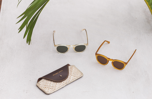 Maison Mata woven pandan and leather glasses case beside two round acetate frames on pale concrete