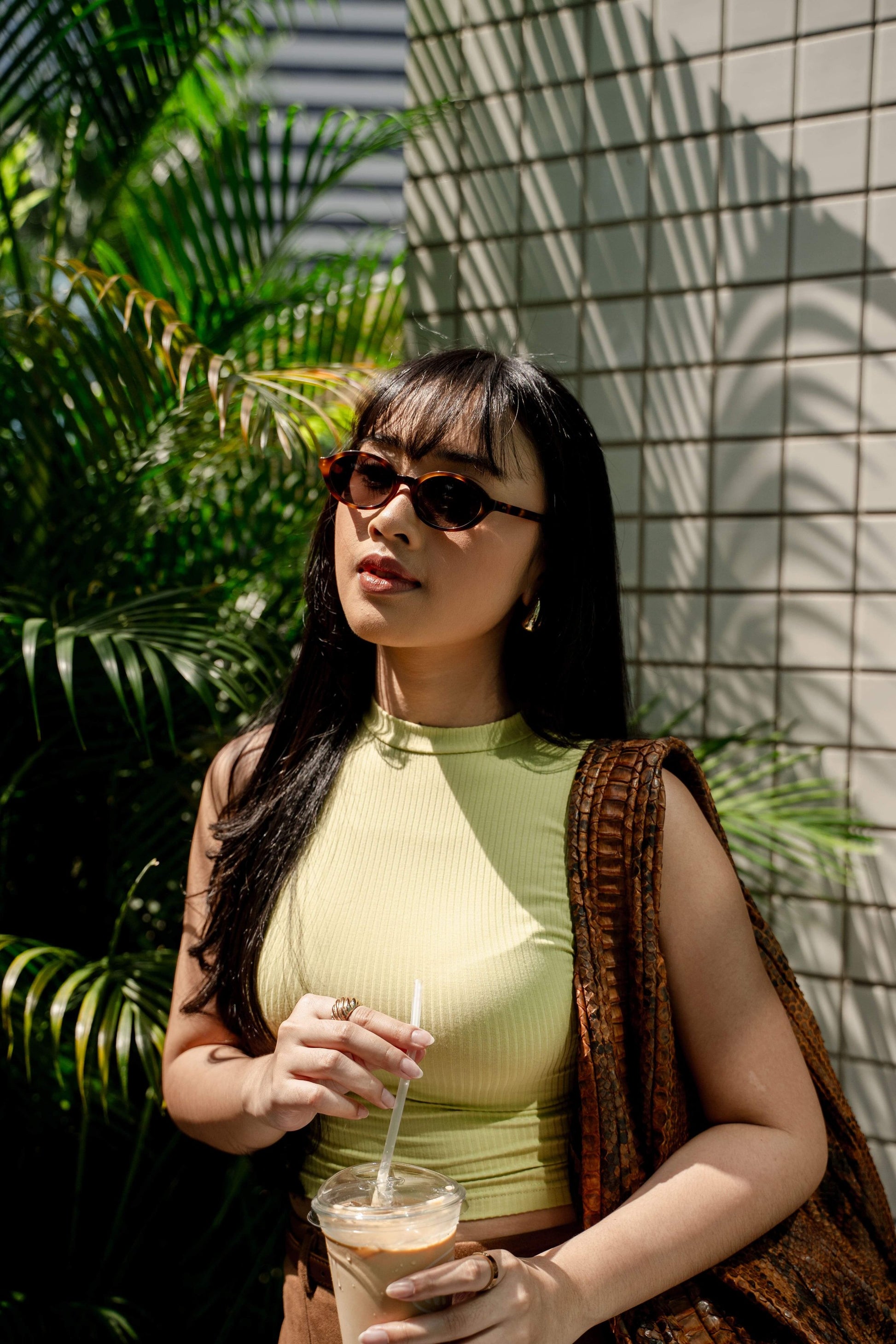 Woman in Cove Caramel Swirl oval sunglasses holding an iced coffee under palms in Bali