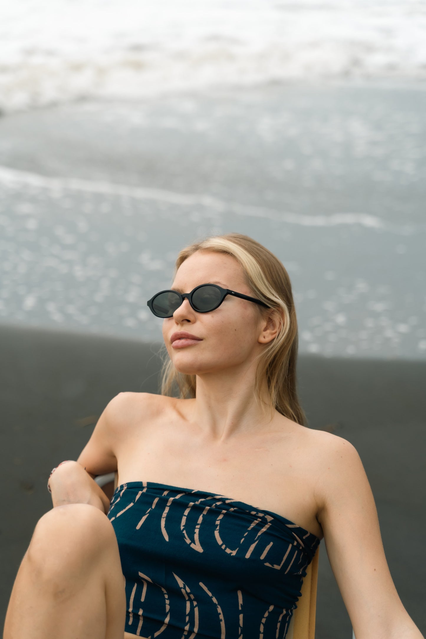 امرأة ترتدي نظارة شمسية بيضاوية سوداء Cove بلون Onyx Night جالسة على شاطئ رملي أسود في بالي.