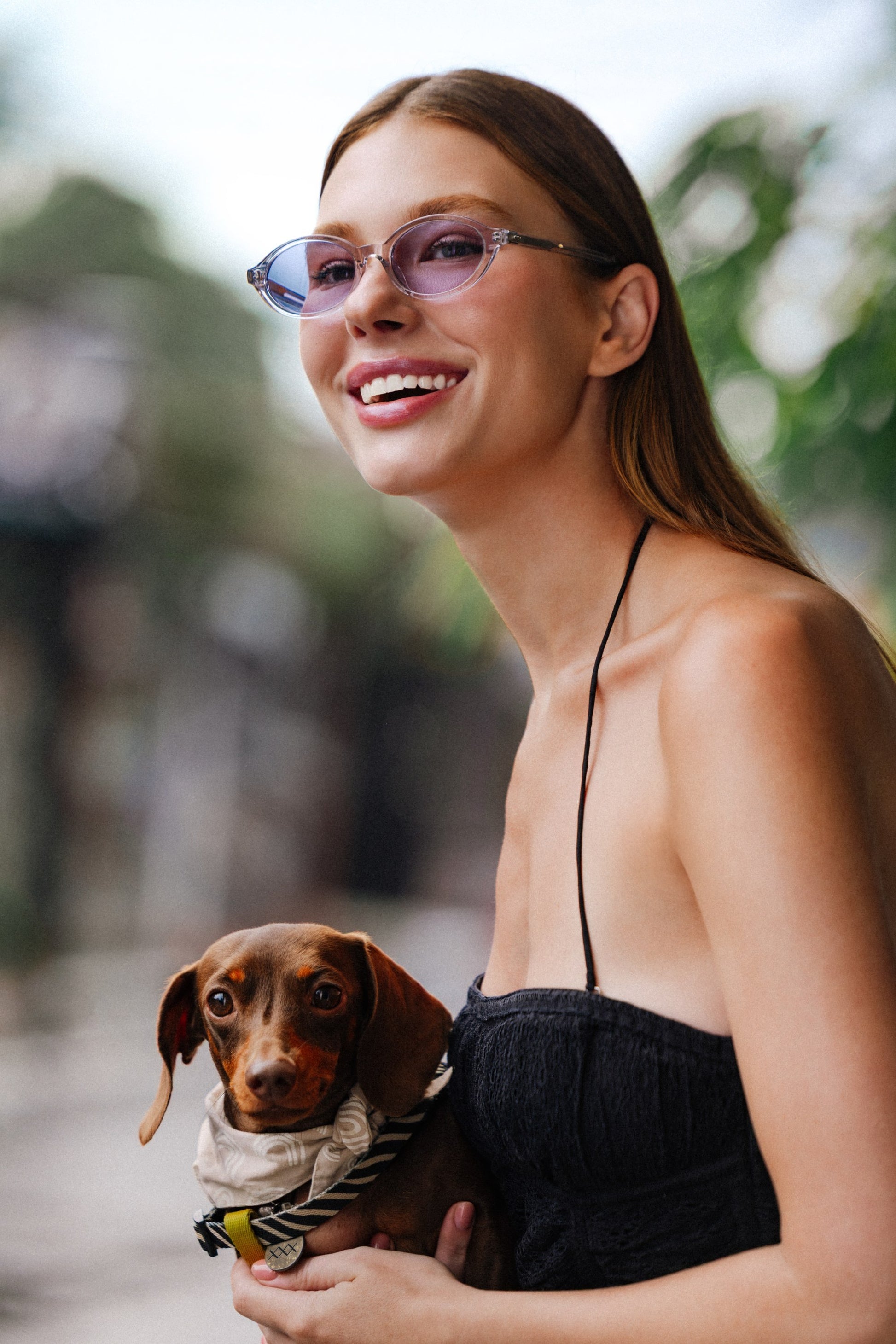 Smiling woman in Cove Silver Mist clear oval sunglasses holding a dachshund puppy
