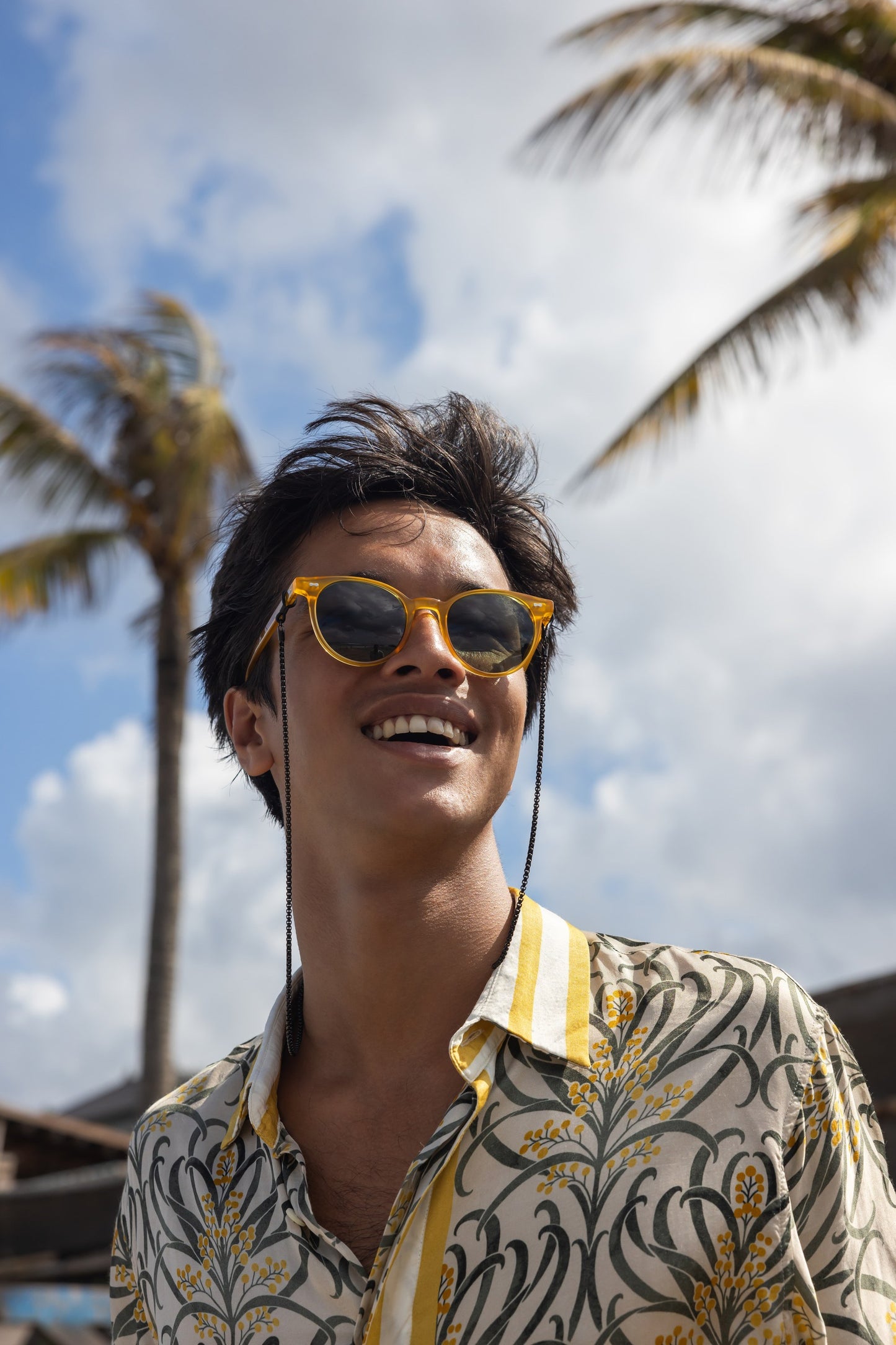رجل يرتدي نظارة شمسية مستديرة Flores Breeze بلون Golden Hour مع سلسلة نظارات تحت أشجار النخيل.
