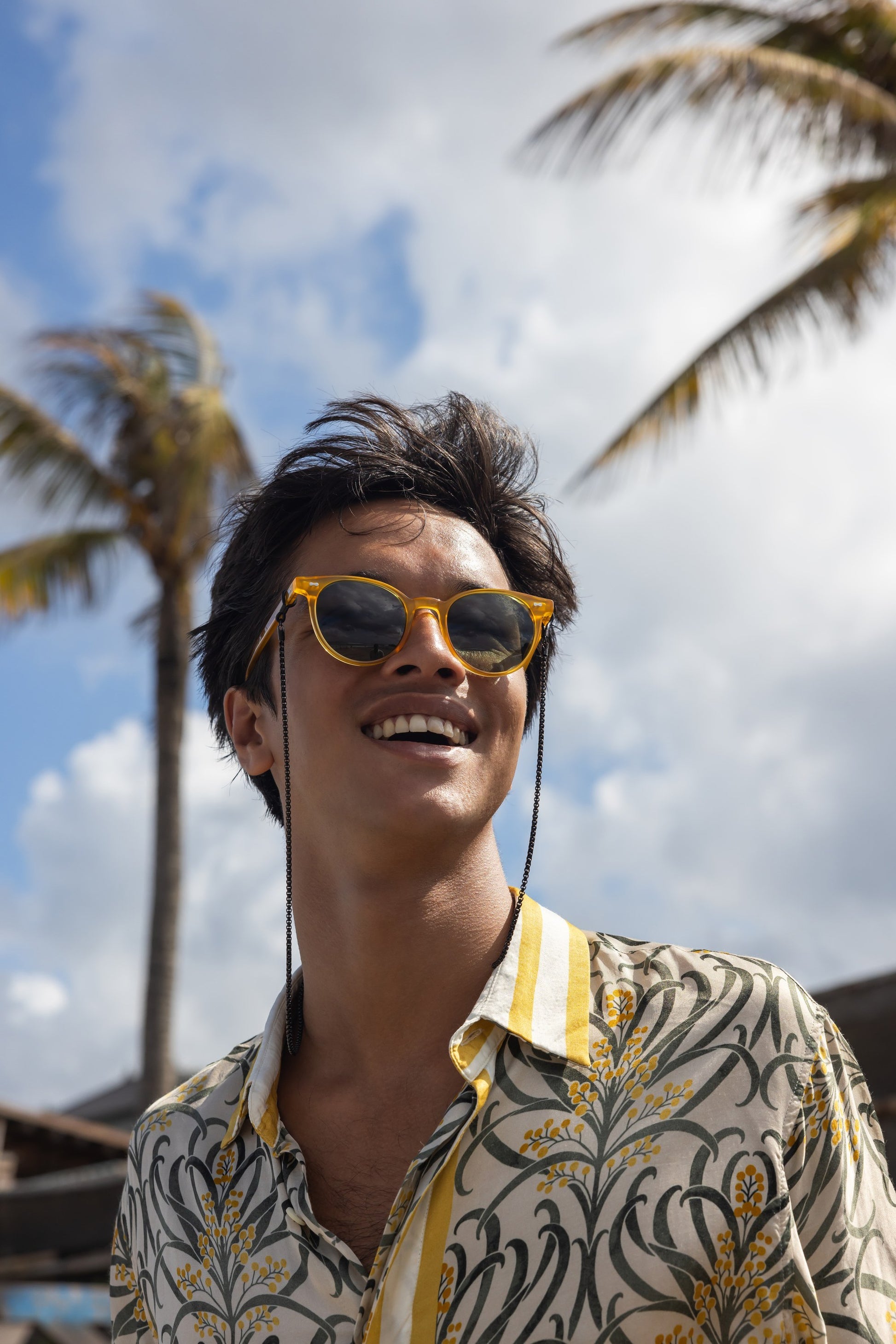 رجل يرتدي نظارة شمسية مستديرة Flores Breeze بلون Golden Hour مع سلسلة نظارات تحت أشجار النخيل.