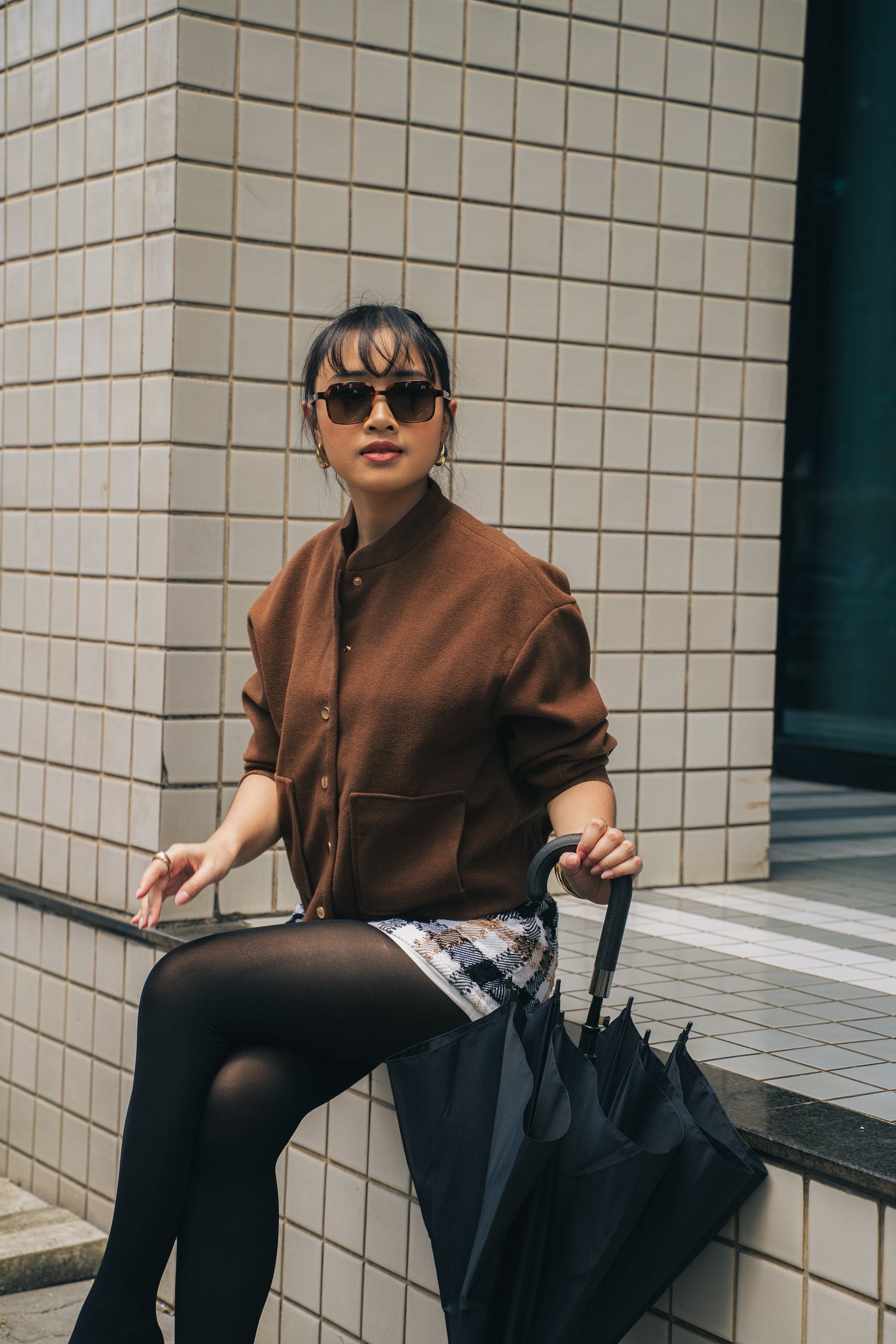 امرأة ترتدي نظارة شمسية مربعة Gili Escape بلون Caramel Swirl وسترة بنية في أحد شوارع المدينة.
