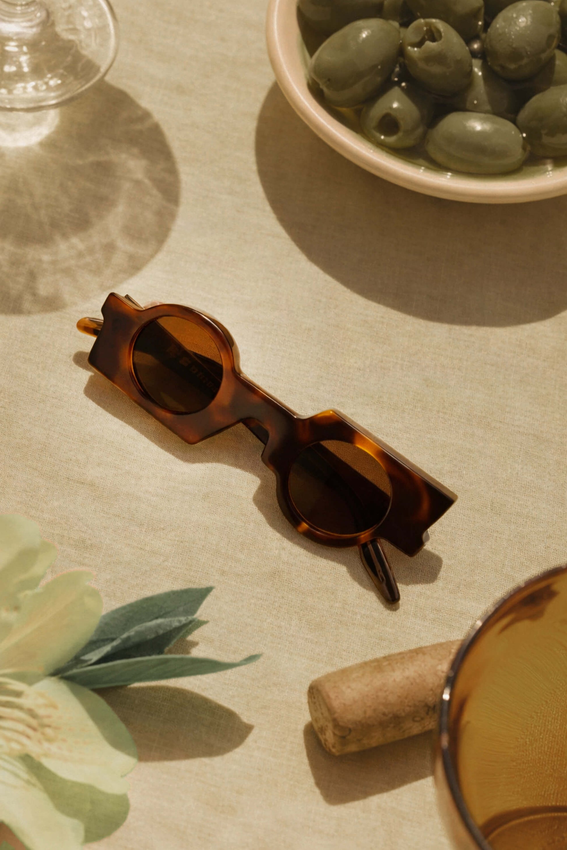 Maison Mata handmade tortoiseshell acetate geometric sunglasses on a linen table with olives and wine — lunettes de soleil géométriques acétate écaille sur table en lin — kacamata geometris asetat tortoise di meja linen — gafas geométricas carey sobre mesa de lino — Bali handmade eyewear